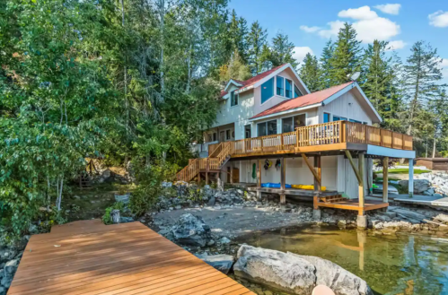 A scenic lakeside house with a wooden deck, surrounded by trees and rocky shoreline under a clear blue sky.